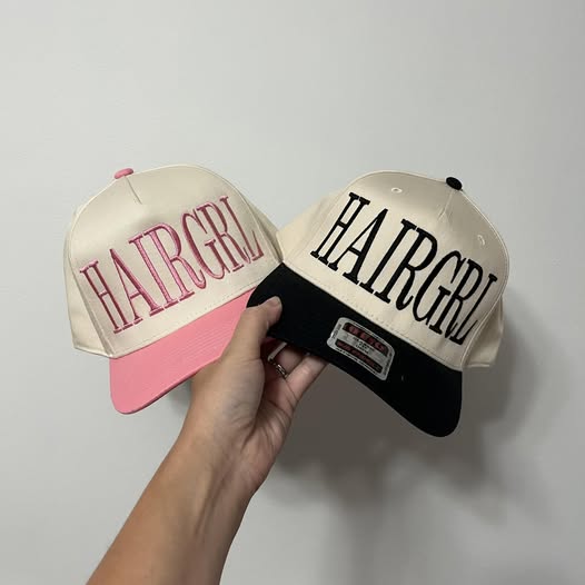 Two hats with 'HAIRGIRL' text, one pink and one black, held against a gray background.